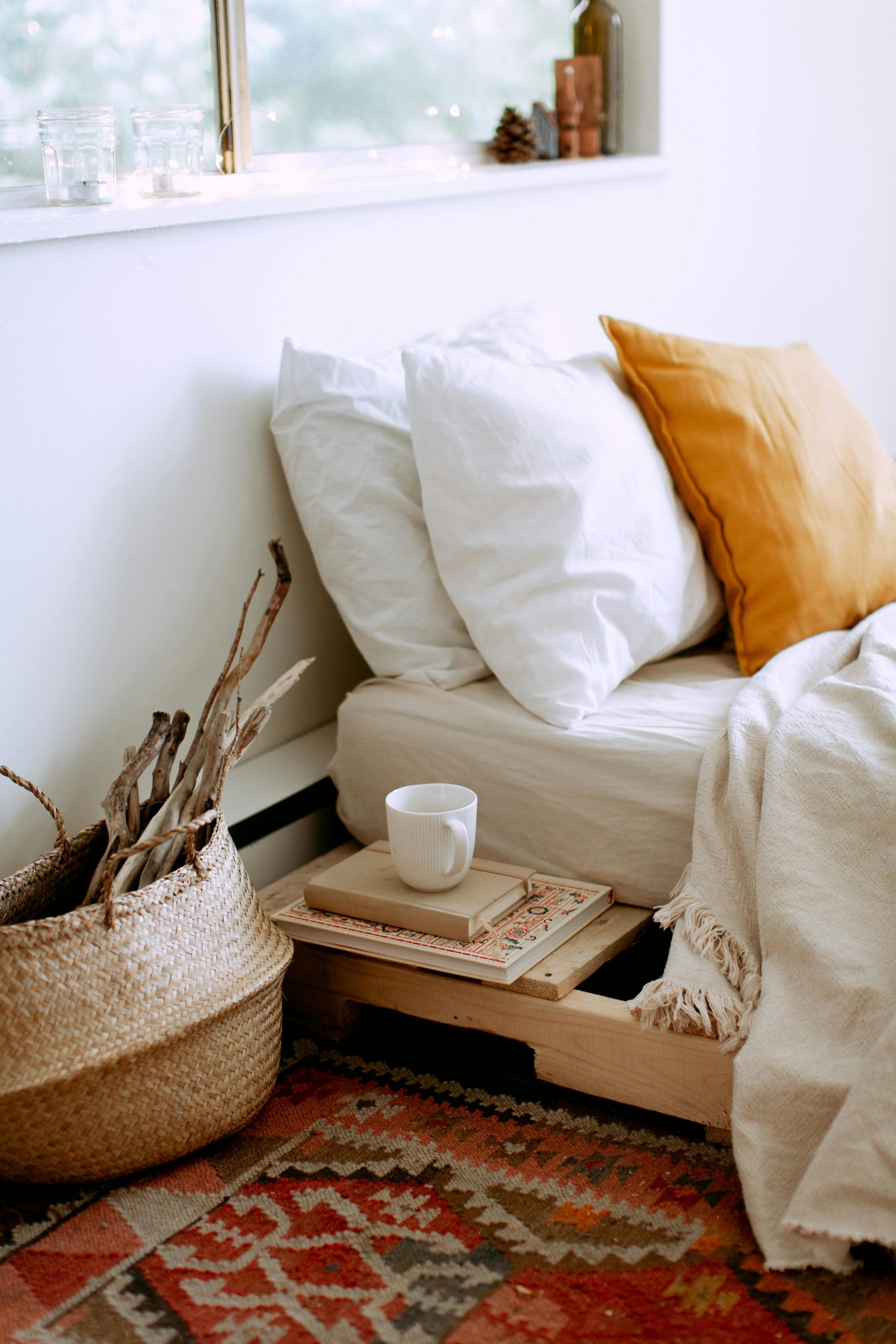 Stylish cozy bedroom interior with bed located near window with white linen, cushions, tray with cup of coffee, basket with dry wooden branches and carpet on floor